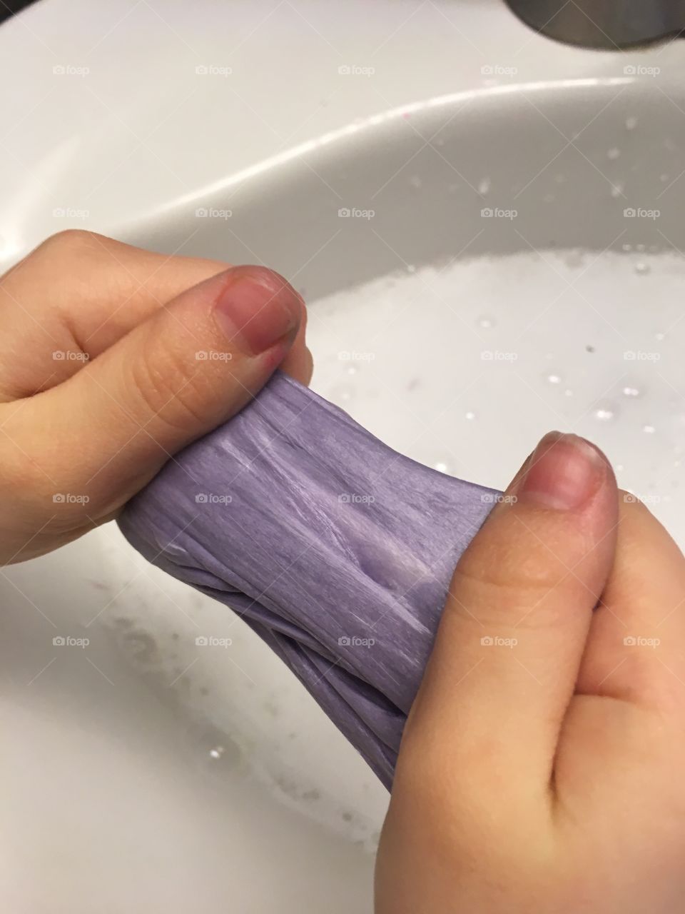 Child making homemade purple slime over a white sink filled with soapy suds and bubbles. 