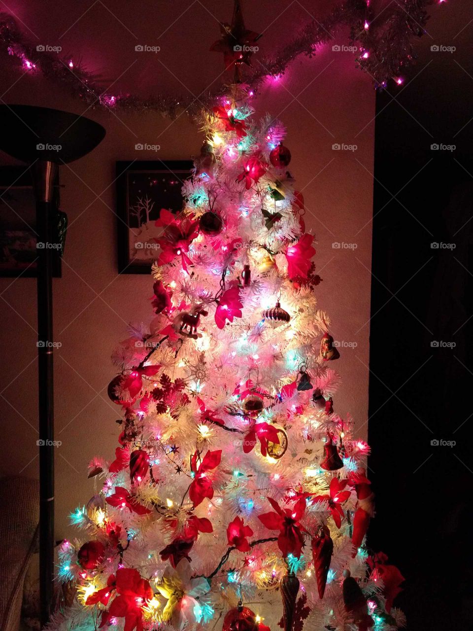 White Christmas tree radiates a warm pinkish glow amid bright poinsettia lights and quaint brass ornaments.