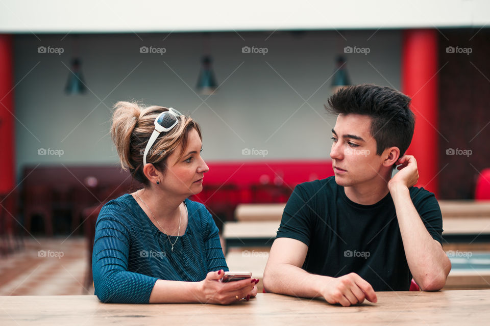 Woman and young man talking together and using mobile phones sitting by a table in cafe