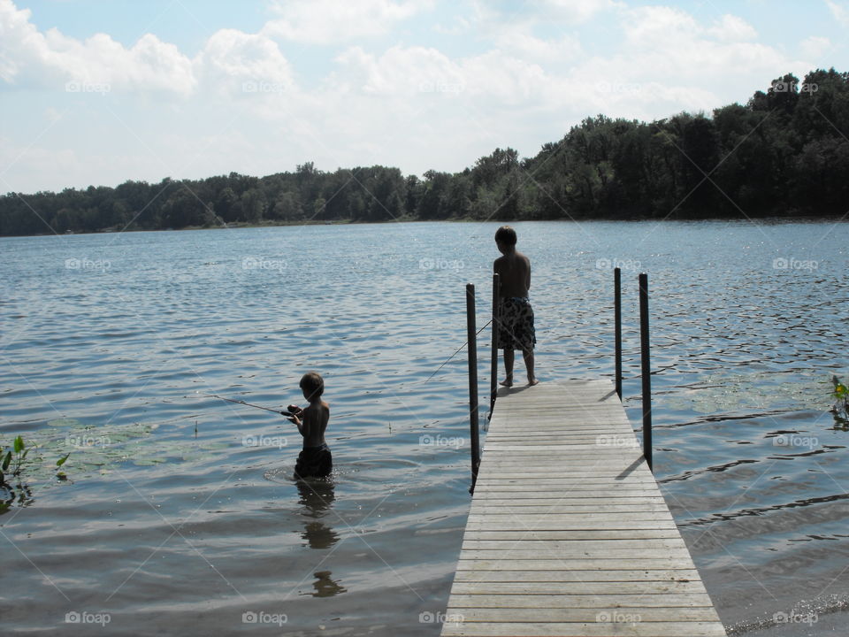 Two boys fishing at a lake. One in the water and one out.