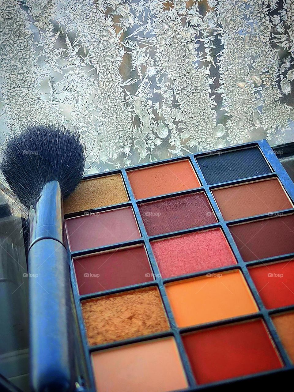 A palette of multi-colored eye shadows and a black brush on the background of a frozen window with a winter pattern