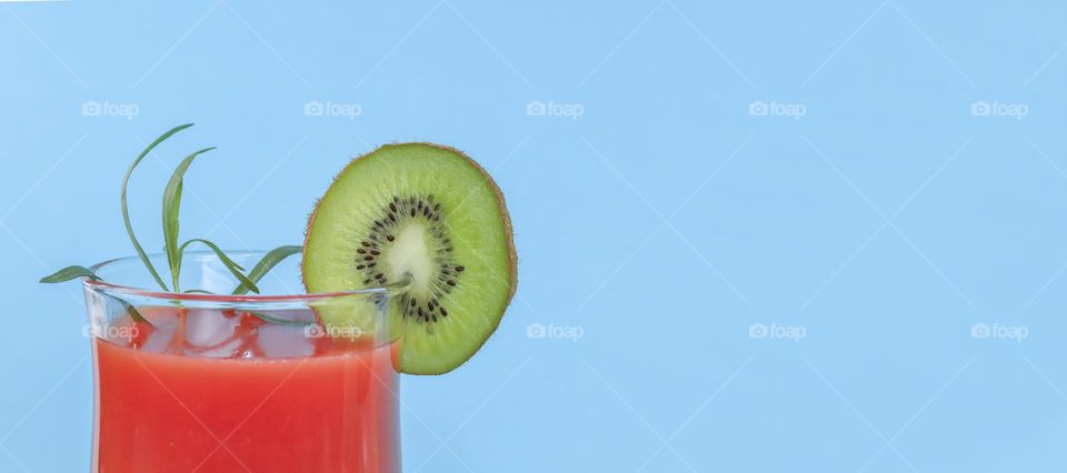 One glass of red watermelon smoothie with a round piece of green kiwi fruit and a sprig of rosemary stands on the left against a blue background with copy space on the right, close-up side view. Summer drinks concept.