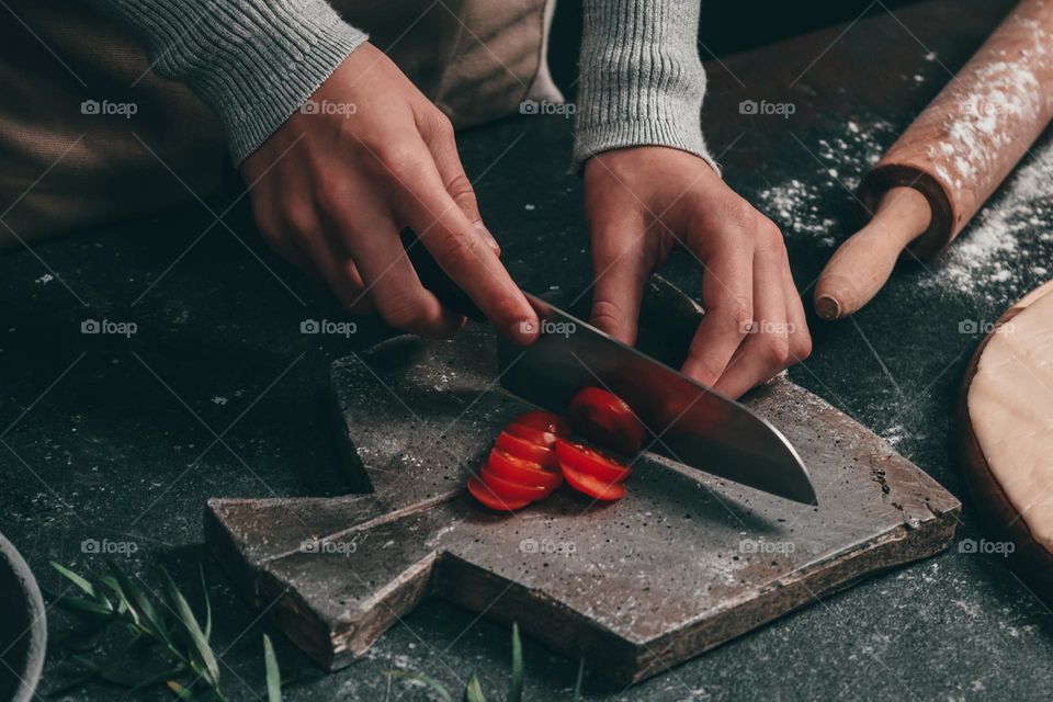 The hands of a young caucasian teenager girl cut red tomatoes with a knife for making pizza while standing at a dark stone table, close-up side view. Home cooking concept.