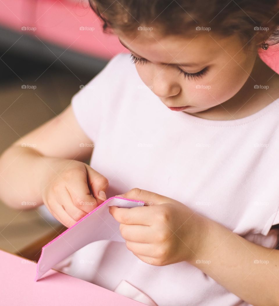 A beautiful little caucasian girl peels off a fake sticker while sitting at a children's table in her room, close-up side view. Concept for creative kids, needlework, diy, easter preparation.