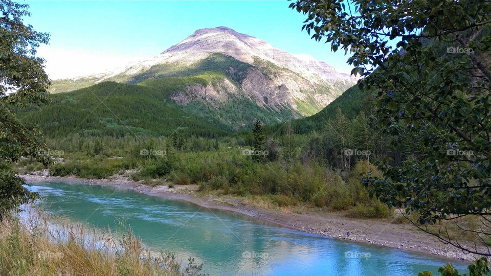 Aqua green waters of the Toad River flow beneath awe-inspiring mountains in Canada on a sky blue summer day.