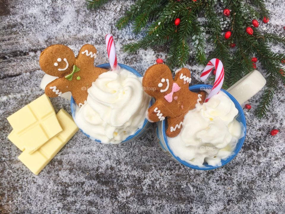 Two mugs of white chocolate with whipped cream, gingerbread man and candy canes on a snowy table with pieces of white chocolate and evergreen branches beside