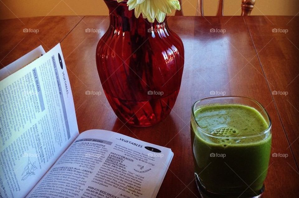 Fresh green smoothie. Table, with red vas of sunflowers, vegetable seed catalogue, book. 