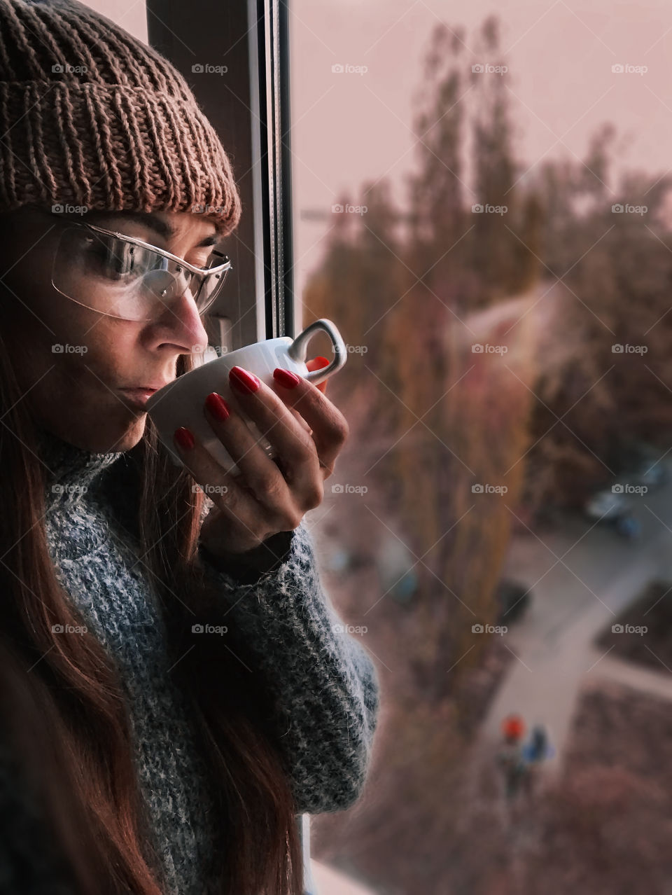 Young woman in knitted sweater enjoying hot tea/coffee at the window 