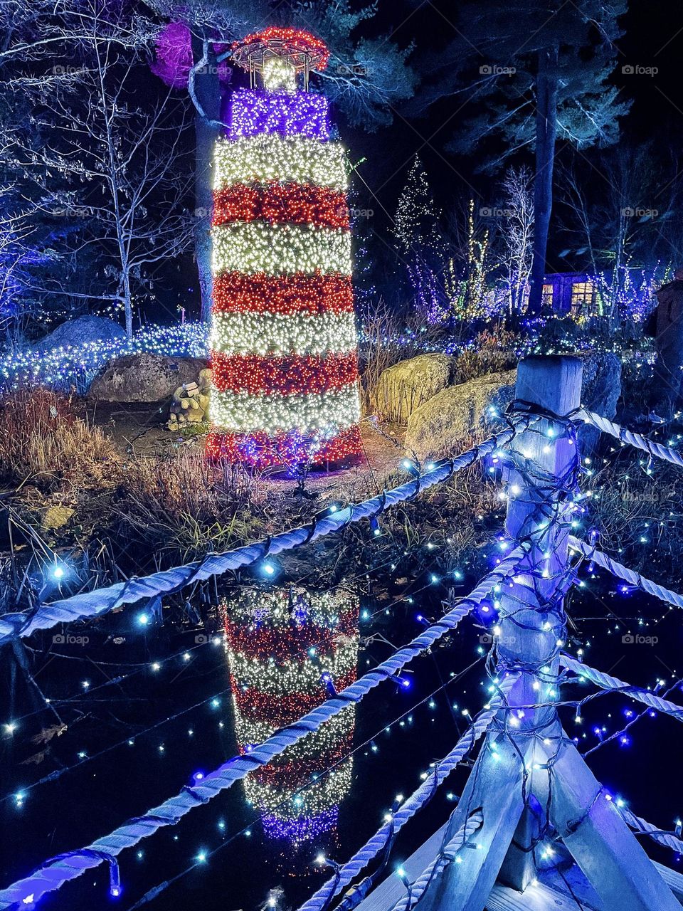 A lighthouse made of multicolored lights reflects over a pond.