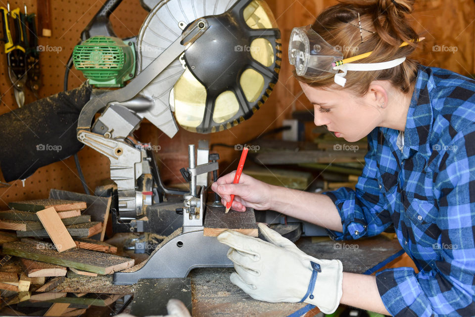 Woman wearing safety goggles and gloves while cutting wood with a miter saw in a shed