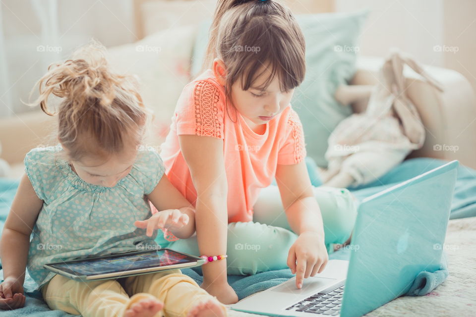 Little sisters with gadgets(laptop and tablet) in the bed.