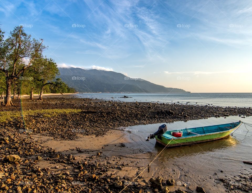 A small boat sits in a shallow sea at low tide with tree lined hill back ground. 