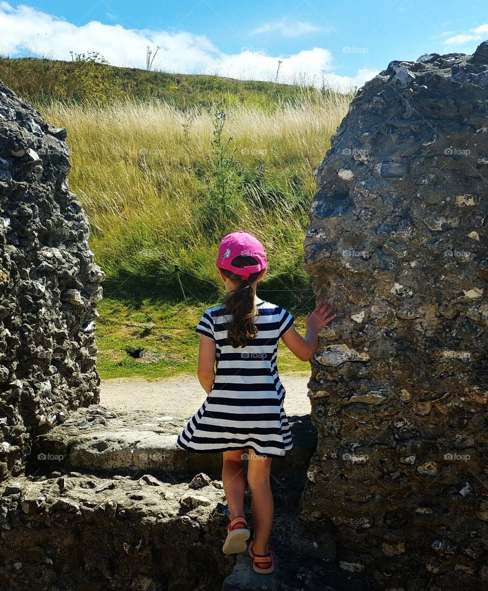 little girl stood by ruined wall in bright sunshine looking out towards feild
