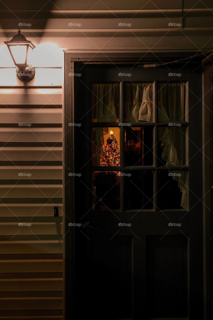 View of a festive holiday tree through the rectangular window panes of a house door at nighttime with the porch light on. 