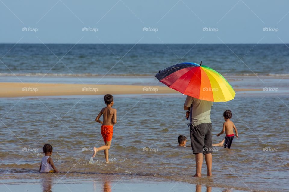 A father with a rainbow umbrella shields from the sun as he watches his children play in the sea. 