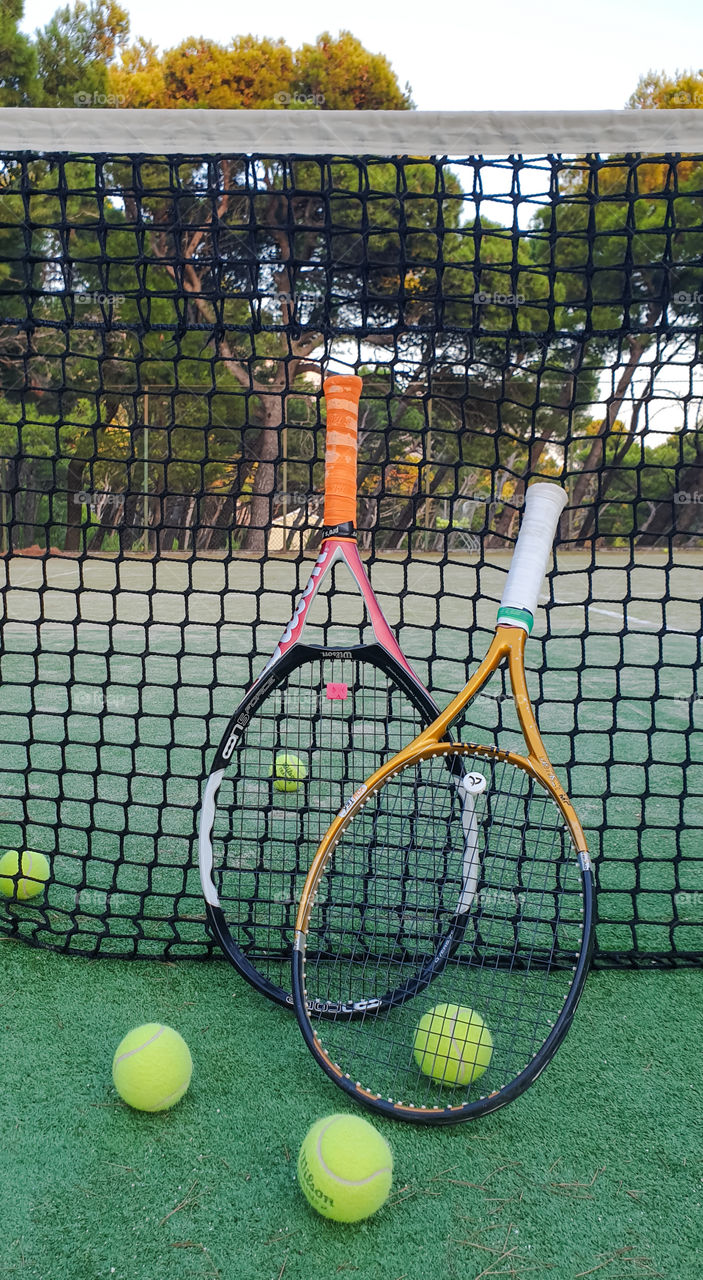 Attributes of the game of tennis. Two tennis rackets and tennis balls on an outdoor court in summer near the net against the backdrop of a pine forest and the sea