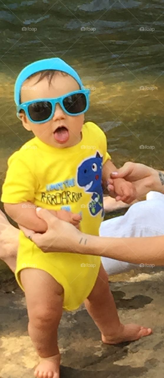 Cute baby in yellow outfit, blue hat,  and blue sunglasses in bare feet and legs with mouth open making this photo adorable.
