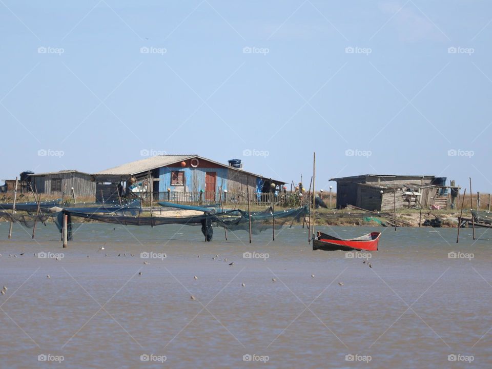Panoramic view of a rustic fishing village with wooden shacks, a vibrant red boat, fishing nets and calm waters under a clear blue sky, evoking a quiet and serene coastal lifestyle.