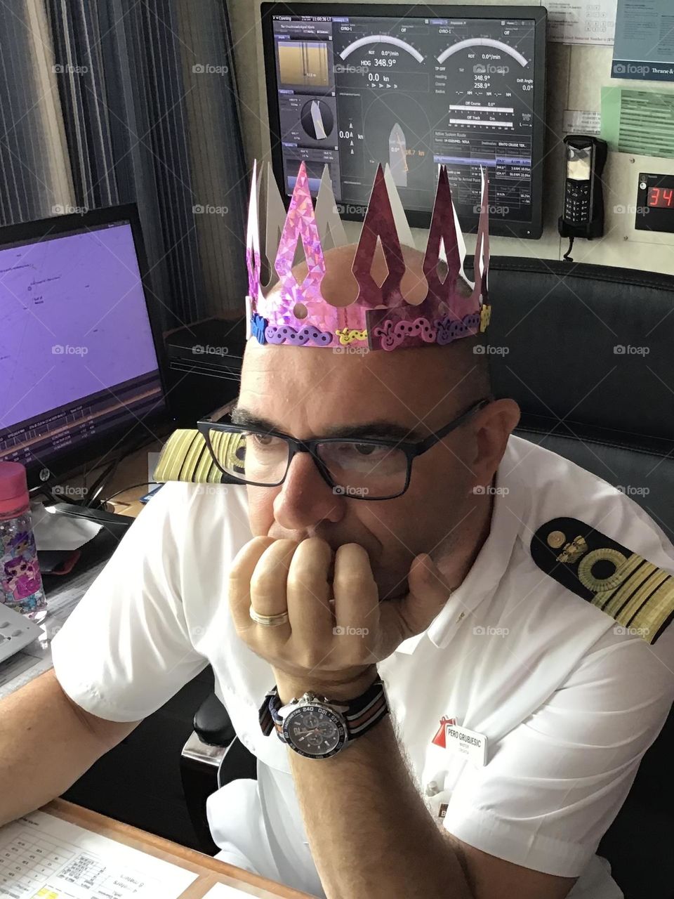 Cruise ship captain proudly wearing a pink sparkly crown made by his 4 year old daughter while working in his office.