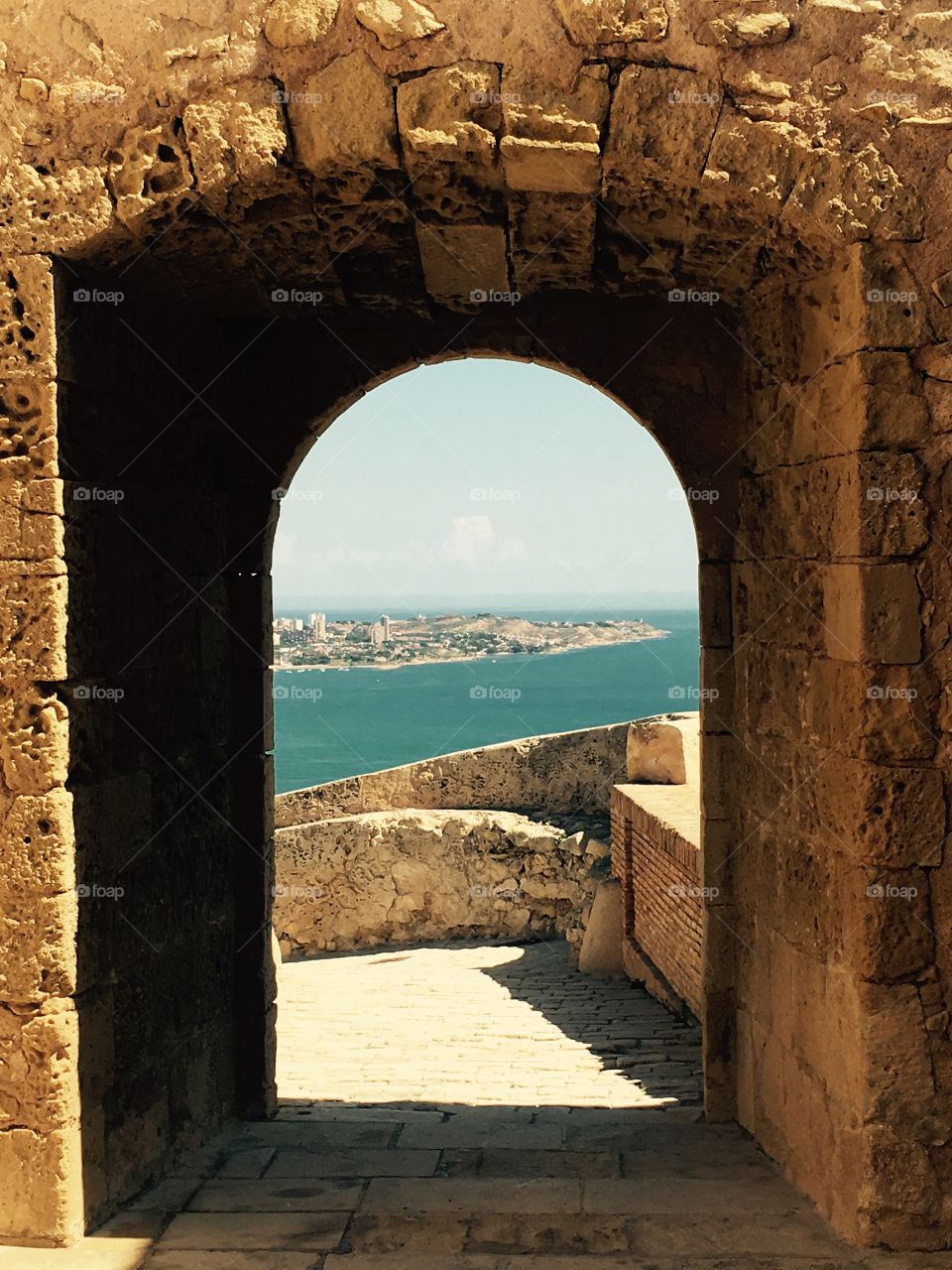 An old brick arch and walkway at Santa Barbara Castle in Alicante, Spain. With a view of the ocean behind it 