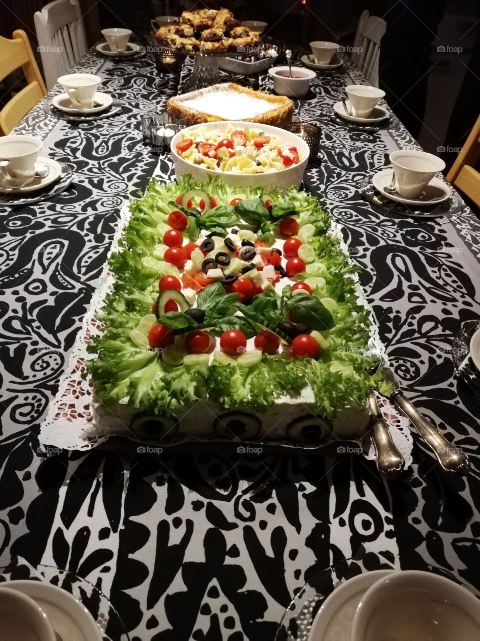 A vegetable sandwich cake on a white doily on a black and white patterned table cloth, salad spoons on the side. Pasta salad, cheese cake, the pieces of chocolate cake and rasperry jam. The light beige coffee cups and tealights in the glass bowls.