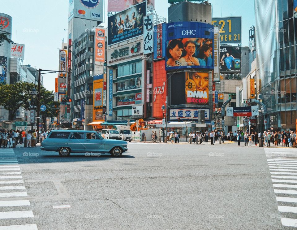 There is a blue car driving through an intersection in busy Tokyo. It is a clear day and the photo is 1970’s themed. 