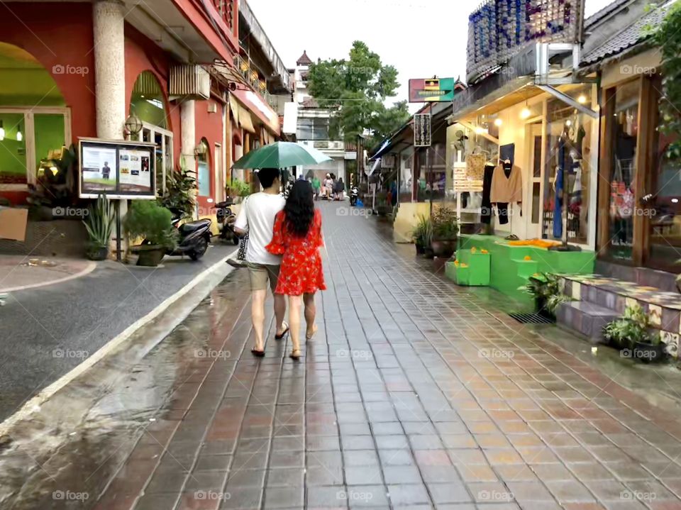 Walking in stride 2. Adult couple with open umbrella. Brick paved street. Shops. Ubud, BalI. 2018.