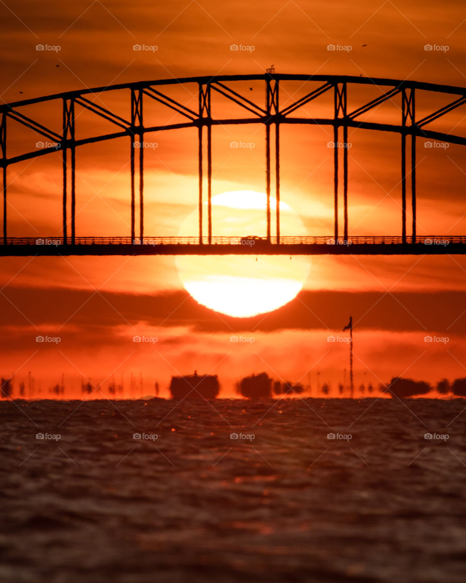 Silhouette of a car driving over a steel tied arch bridge with a large sun setting in the background. 