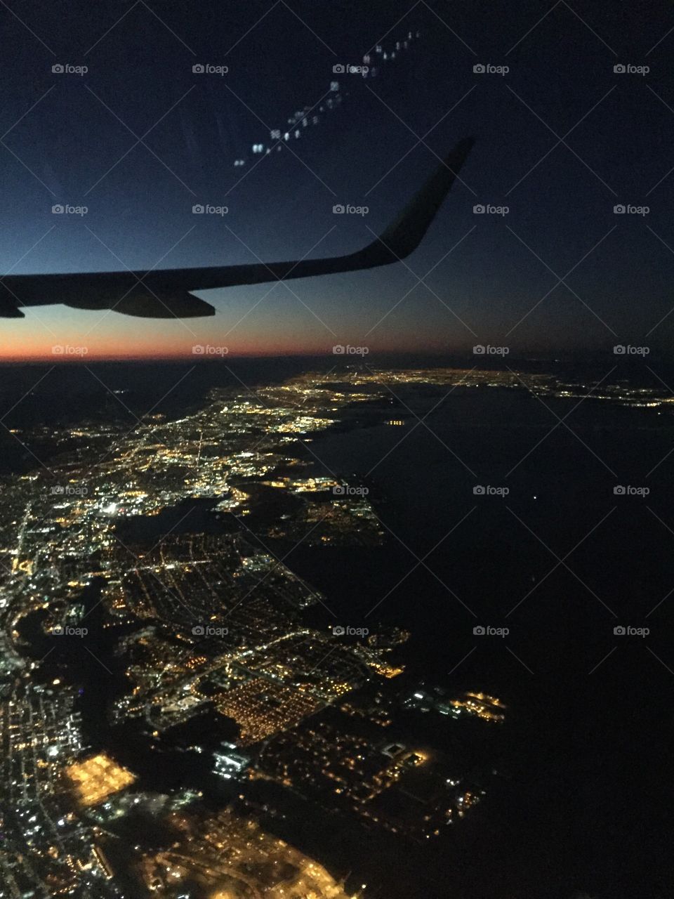 Dusk falls on a city, as seen from a plane. Might be Chicago? The city lights twinkle beneath the deep blue sky 