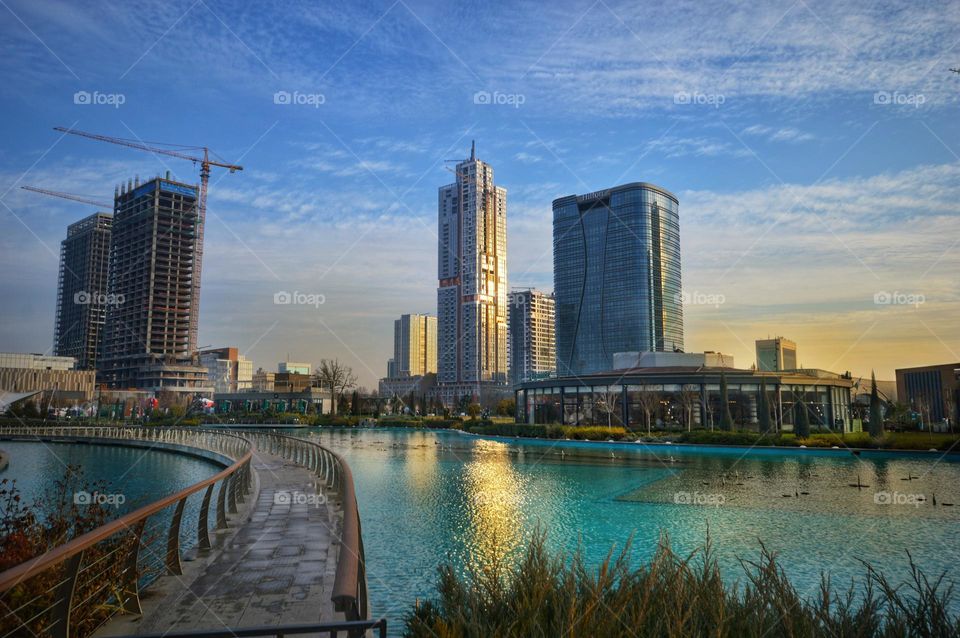 a huge swimming pool with blue water and a bridge in the center of a huge metropolis with skyscrapers and a blue sky at sunset