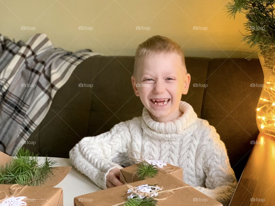 boy in a white sweater decorates gifts with Christmas tree branches, lights