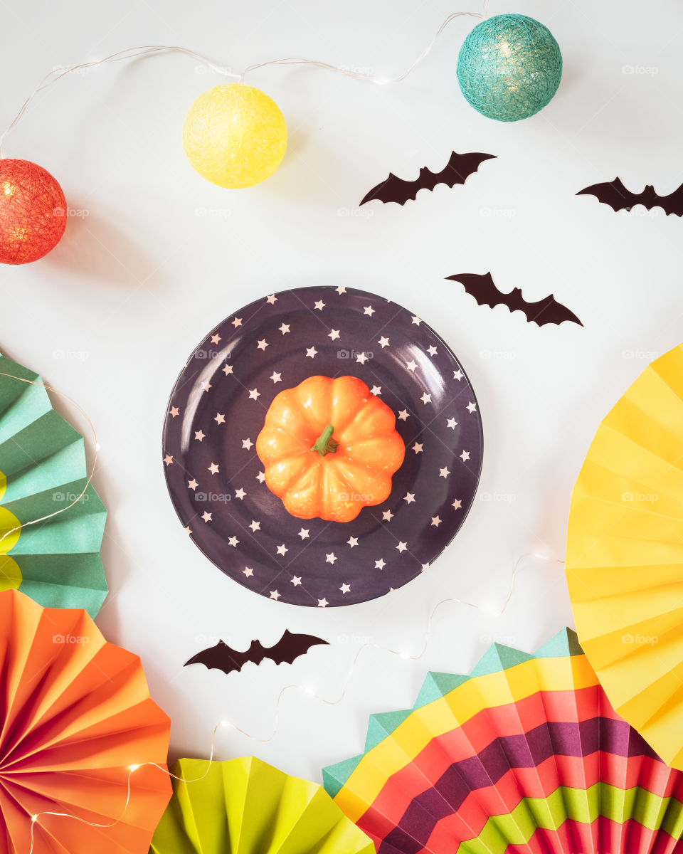 Helloween, autumn composition with small pumpkin on a plate, colorful paper fans, helloween decorations and light garland on a blue background. Cute, fun, Halloween party. Flat lay
