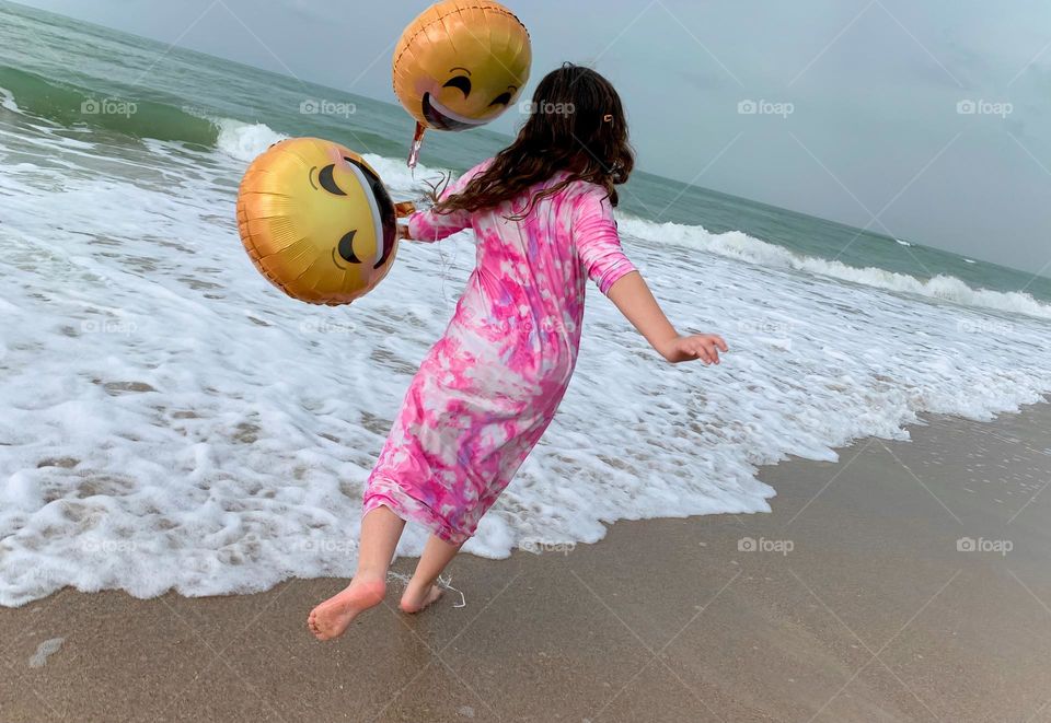 Little Girl Seems To Be Flying And Carried Up In The Air By The Three Helium Emoji’s Balloons On The Beach By The Ocean.