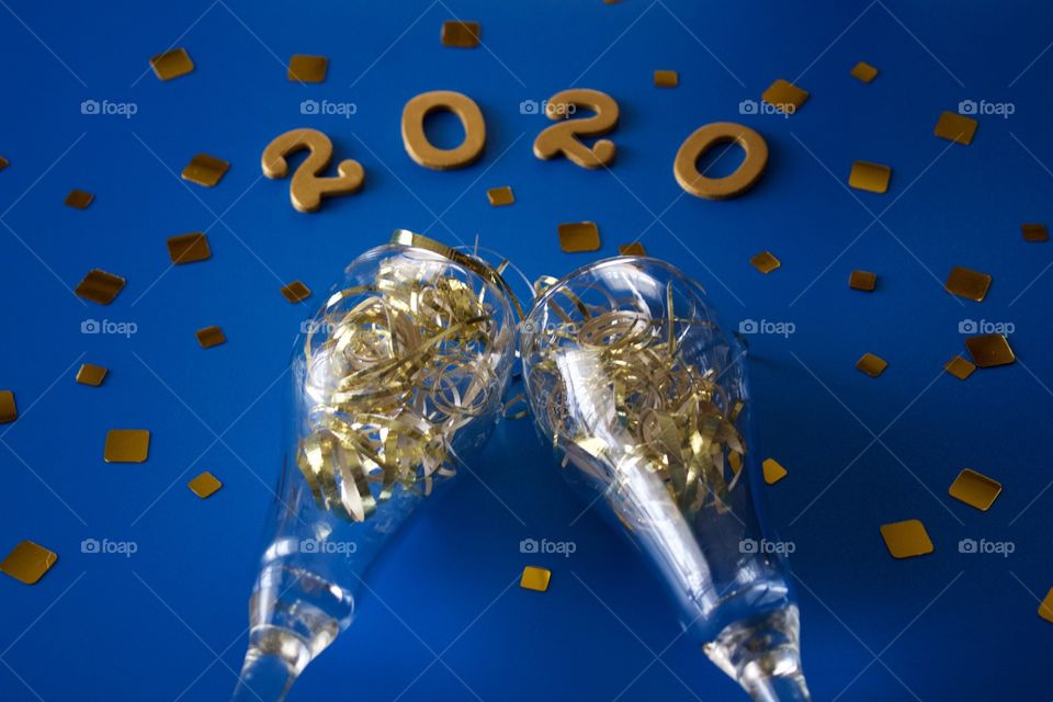 Angled view of a conceptual champagne toast with glittering gold confetti and the numbers ‘2020’ in gold on a classic blue background - landscape view