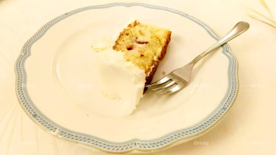 A slice of a wedding cake with cream milk and strawberries served in a white and blue dessert plate with a dessert fork
