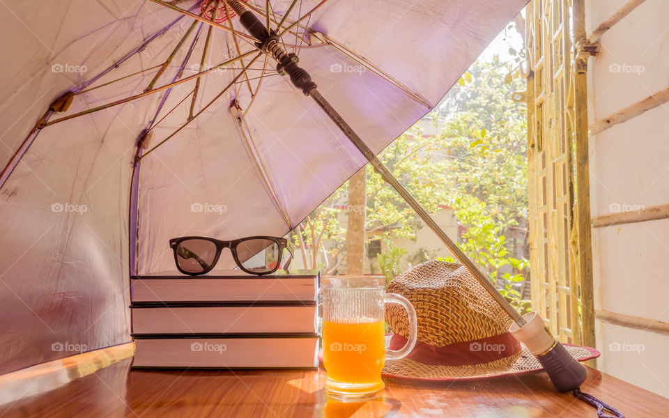 Super Protection from the sun. Sunburn risk concept. Umbrella protecting beautiful woman things. Books, sunglasses, orange juice and a straw hat. Summer holiday concept with copy space room for text.