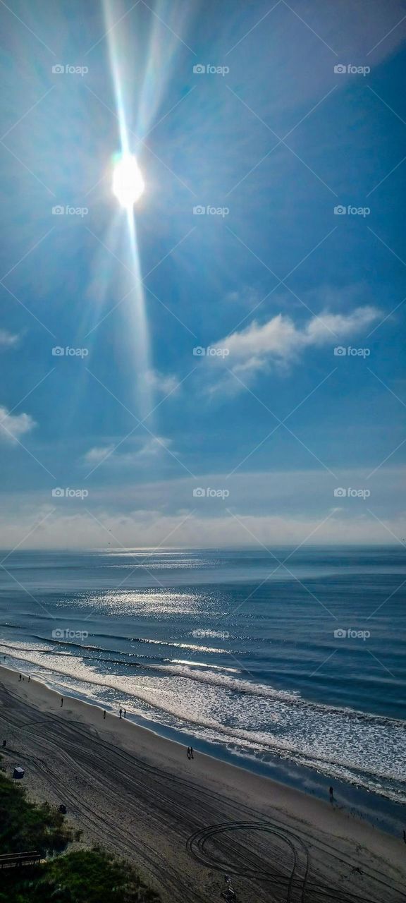 15th floor view of a Myrtle Beach, white stratus clouds and bright bursts of light shining from the sunlight beams making a spotlight on the dark blue water against light cerulean blue skys 