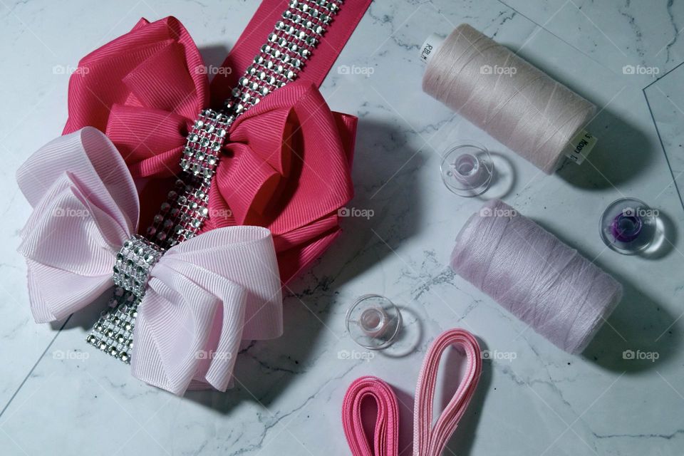 A hot pink & baby pink bow sit on top of bright pink ribbon & silver diamanté’s, accompanied by small & medium sized pink cotton reels & pink ribbon on a simple white marble flatlay background.