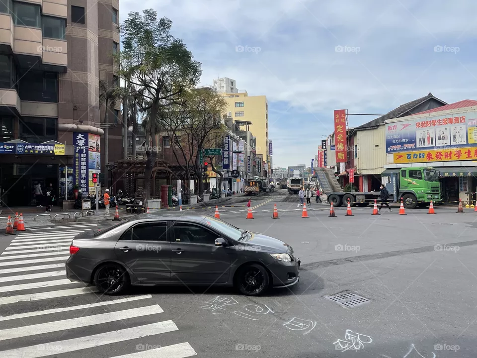 An busy intersection in Taiwan. Across the street is a construction site blocked off with orange pylons. I’m the foreground is a black car turning right. It is partly cloudy outside. 