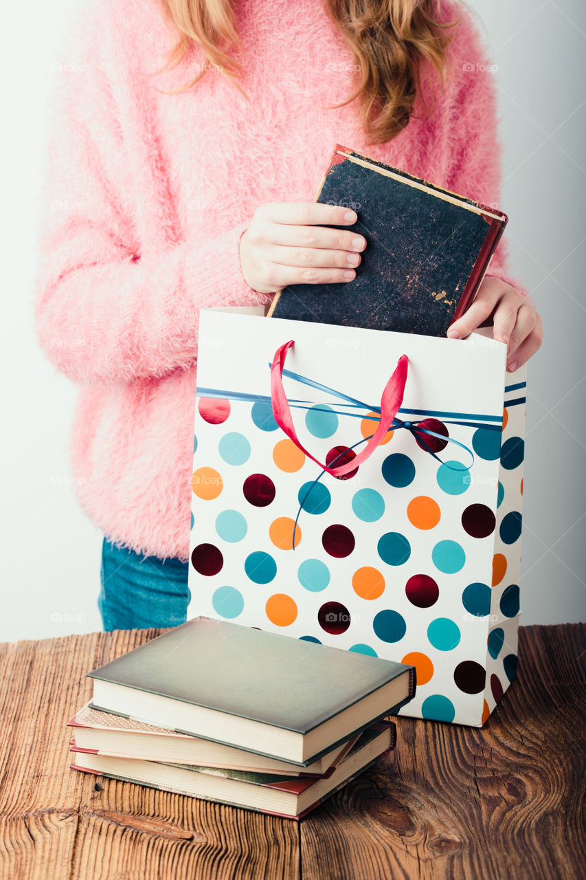 Young girl turning the pages of book in bookstore. Putting the books into paper bag. A few books on a wooden table. Teenager girl wearing pink sweater and blue jeans. Vertical photo