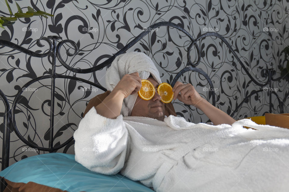 A man and home spa care at home takes care of himself and smears his face with cream, does a manicure at home on a blue background and with oranges and rests.