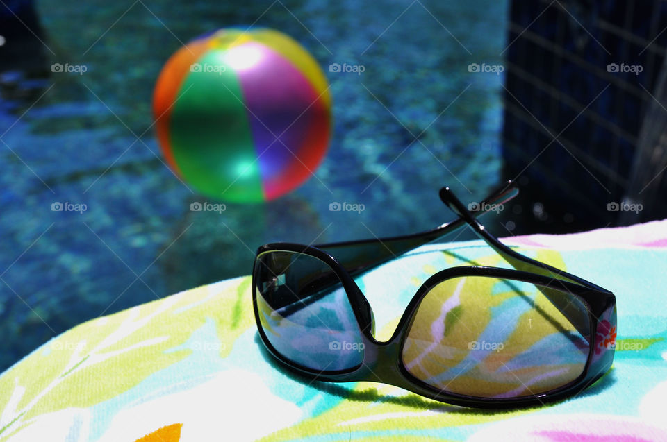 A pair of sunglasses by the swimming pool with a beach ball in the background. 