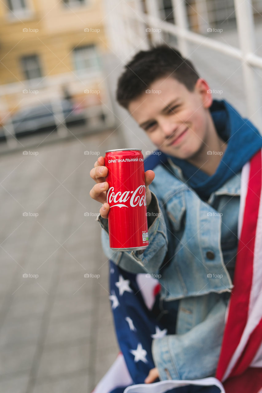 a cute guy, a teenager in denim clothes, holds a Coca-Cola in his hands, drinks it, walks down the street, spring.  stylish boy