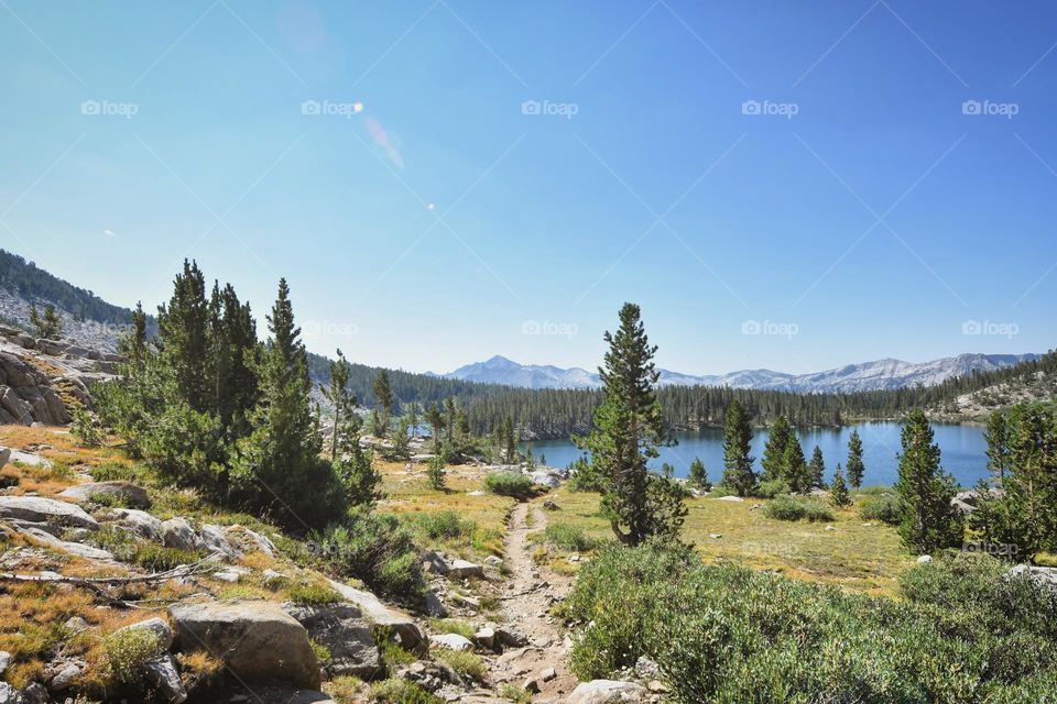 A thin dirt trail wanders down towards a wilderness lake with mountains in the background