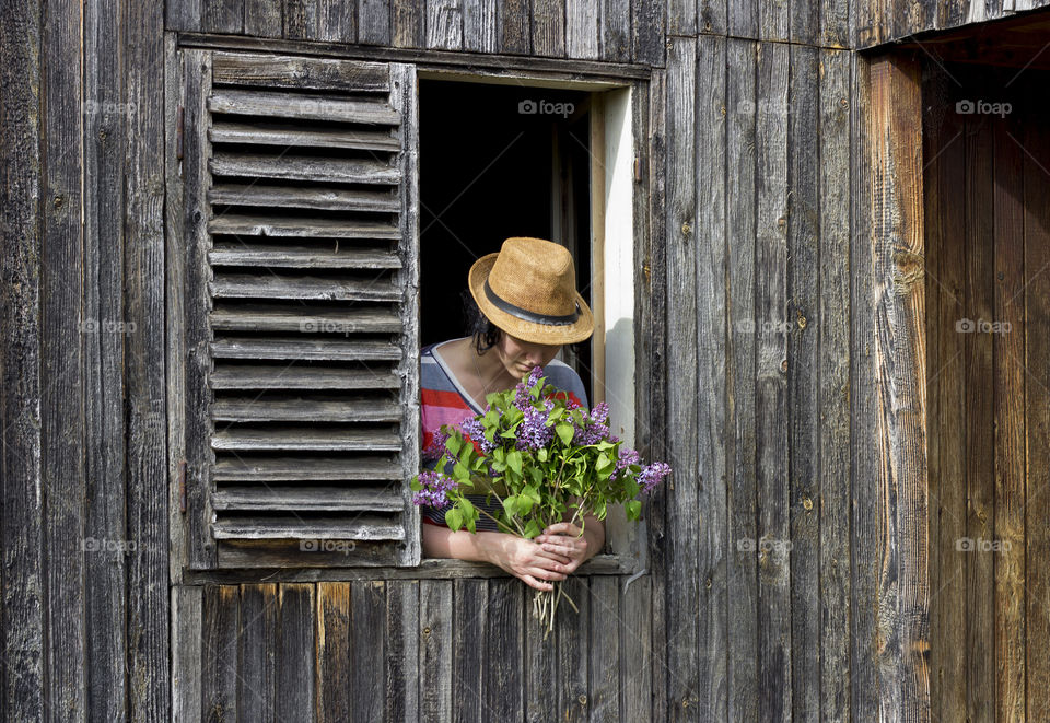 Young woman with a hat holds lilac flowers on the window of wooden country house