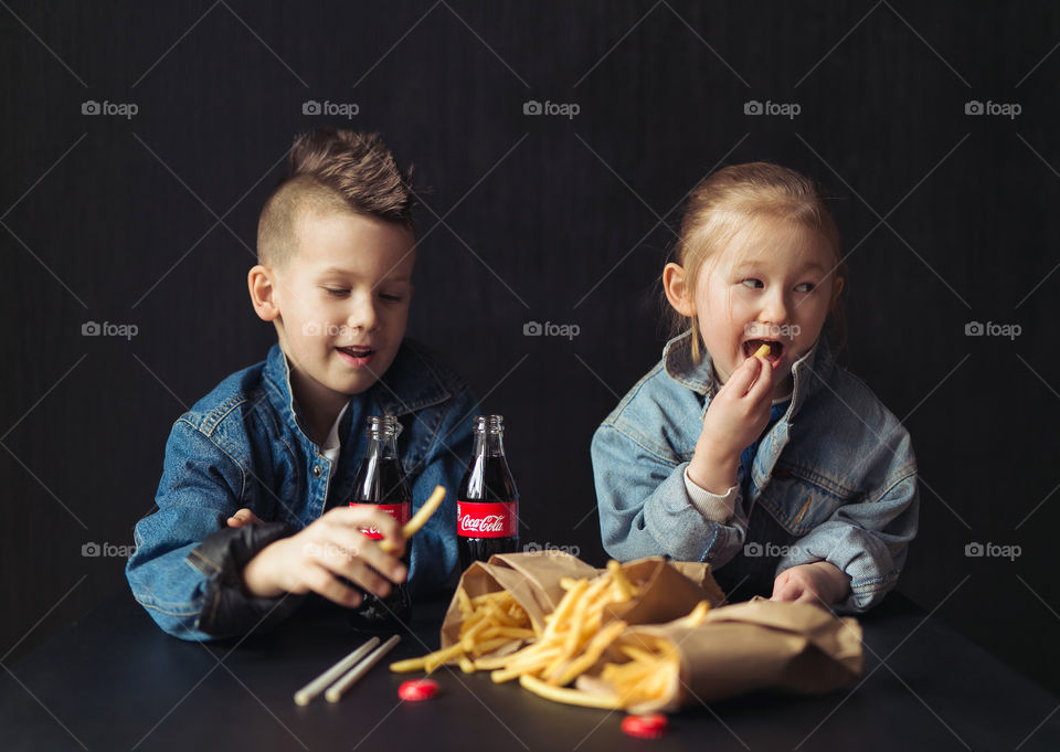 a boy and a girl of seven and five years old, friends, drinking Coca Cola, laughing, having fun, wearing denim clothes. Tasty food on the black table. Lifestyle photo. Happy kids. Emotions 