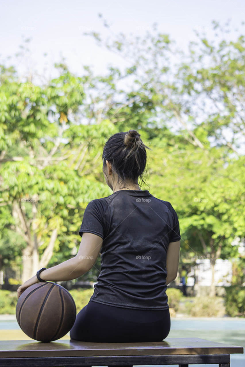 Asian Woman holding basketball sitting on a wooden chair Background blur tree in park.