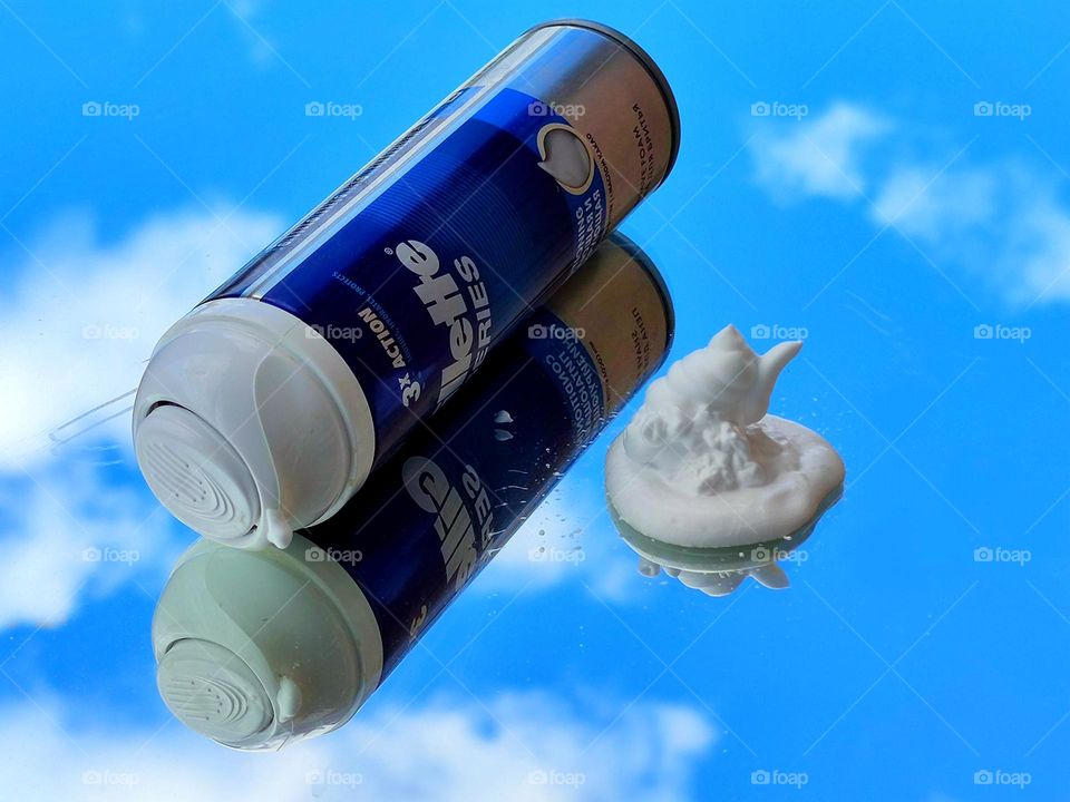 Shaving foam. On the mirror surface there is white shaving foam and a bottle of "Gillette series". A blue sky with white clouds reflects off a mirror surface