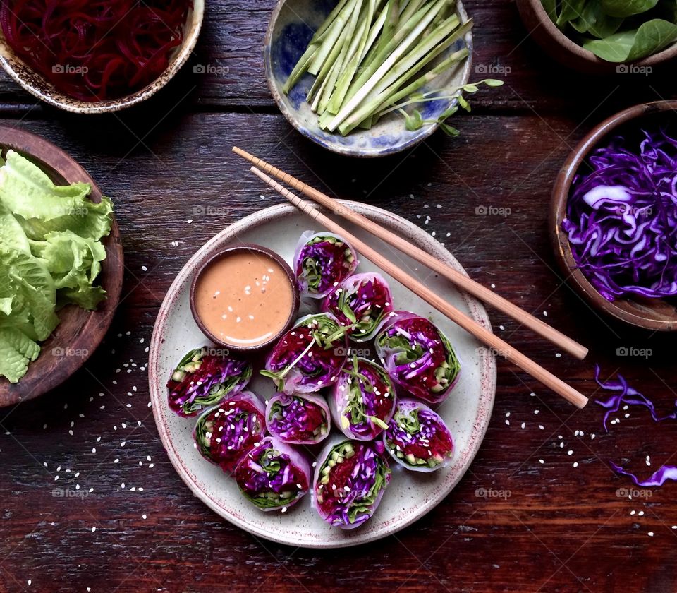 Colorful summer rolls on a plate with dipping sauce and chopsticks next to ingredients on wood table.