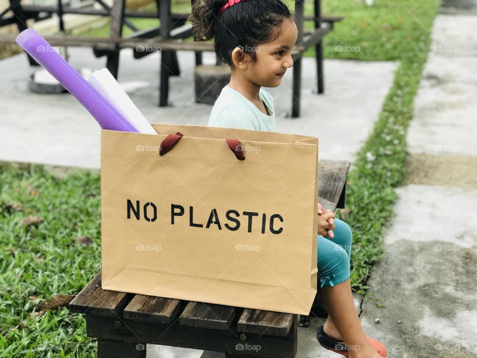 A four year old smiling girl sat on a wooden table and beside her a paper bag which written no plastic on its top.